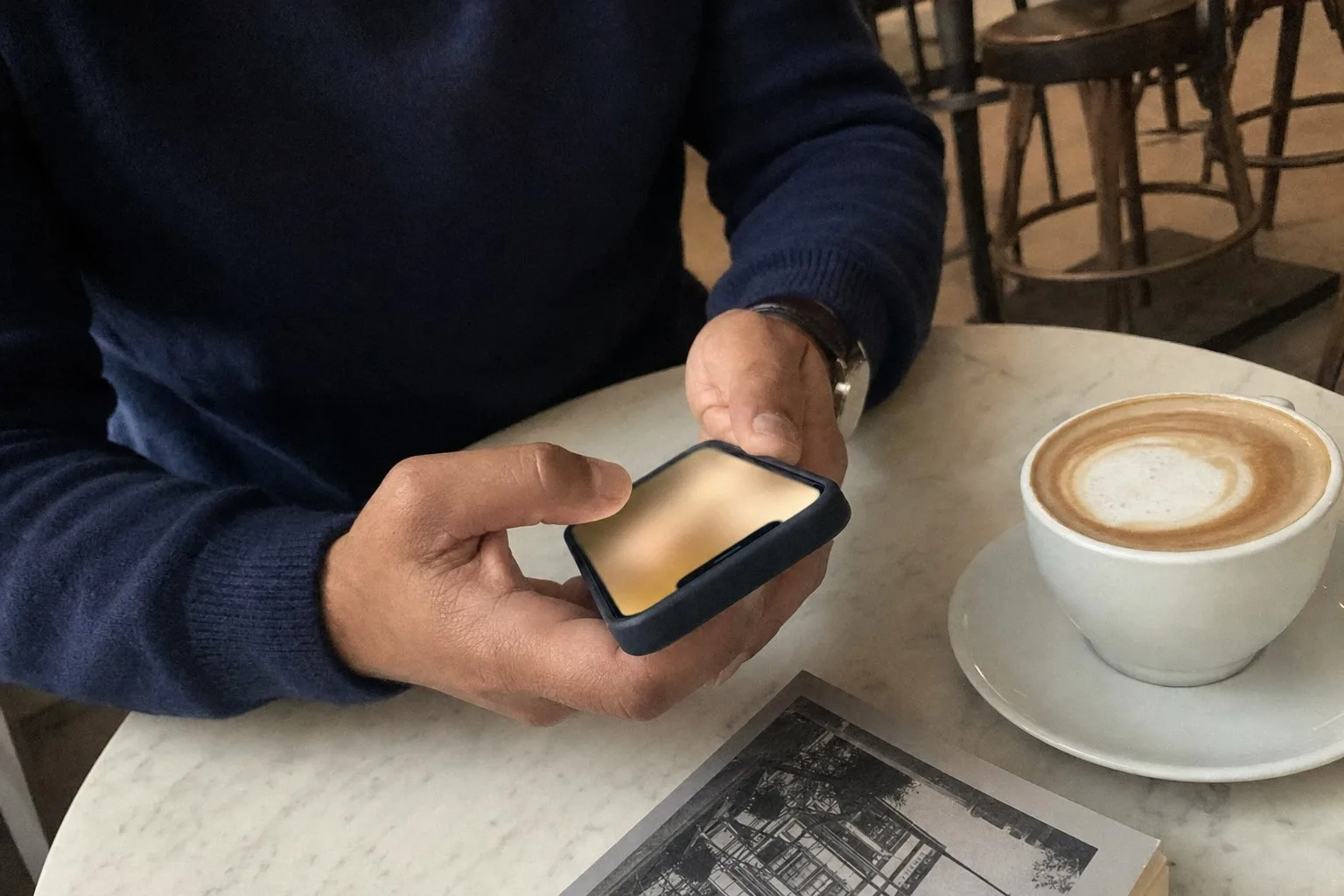 Hands holding a phone on a cafe table beside a coffee and a paperback