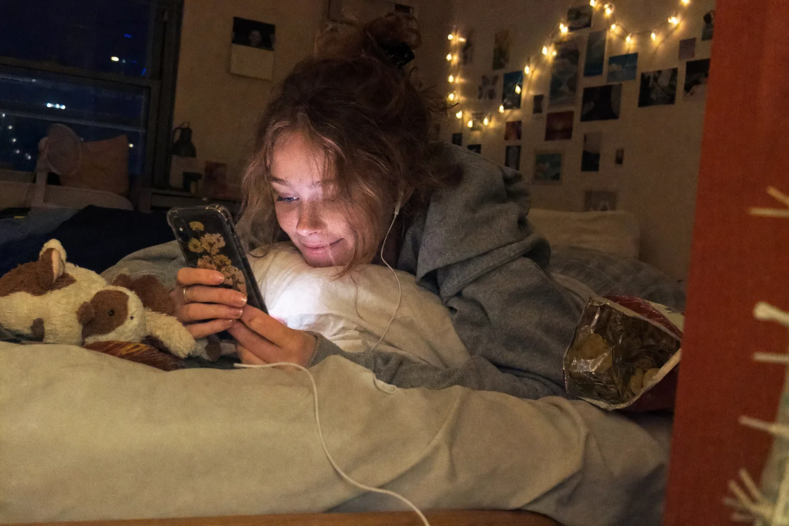 A young woman lying on her bed at night under fairy lights, smiling at her phone with earbuds in
