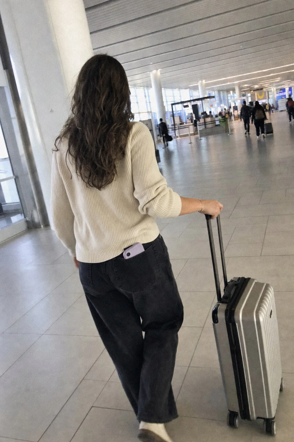 A woman seen from behind pulling a silver carry-on through a bright airport terminal