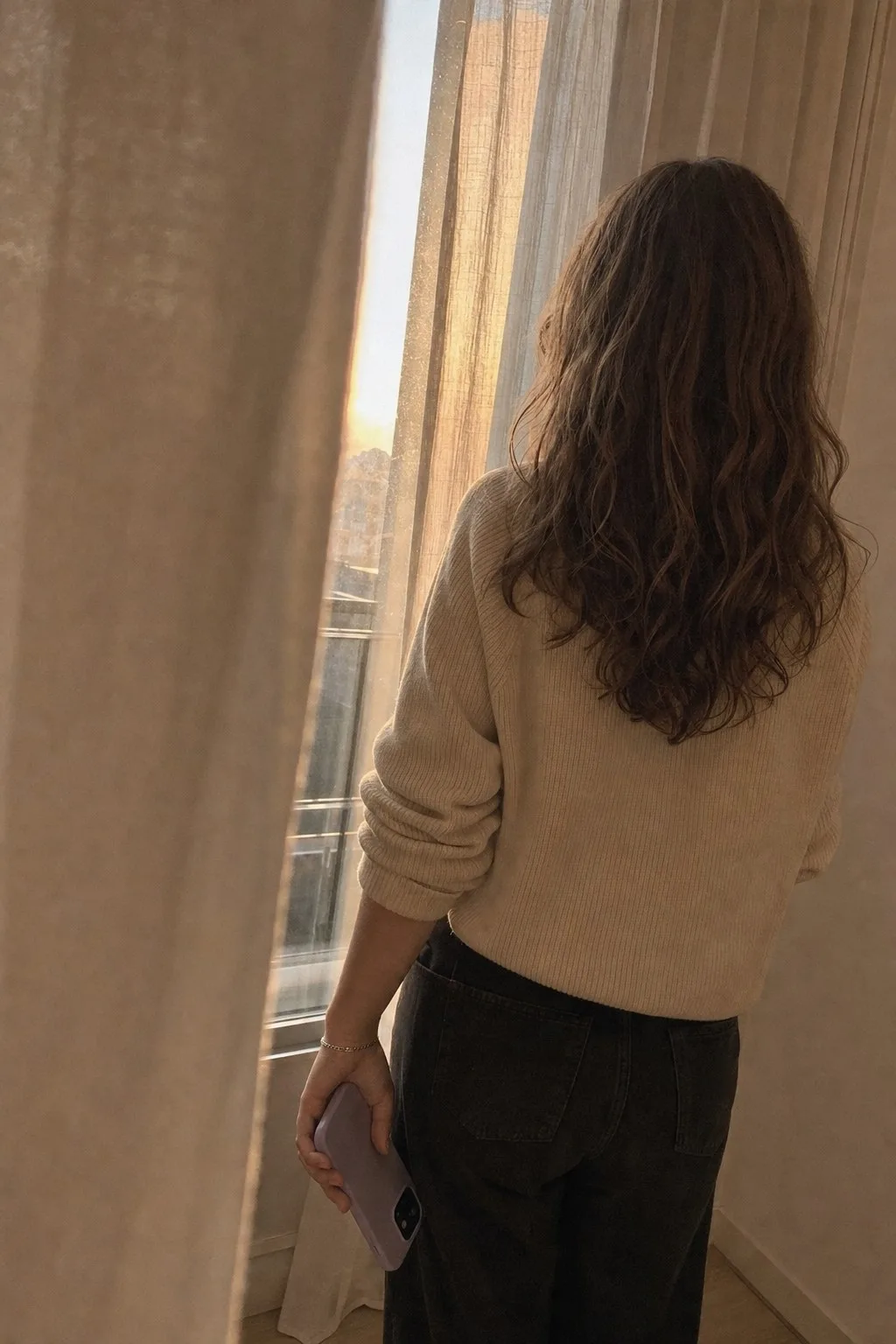 A woman seen from behind at a curtained window in golden light, holding a phone at her side