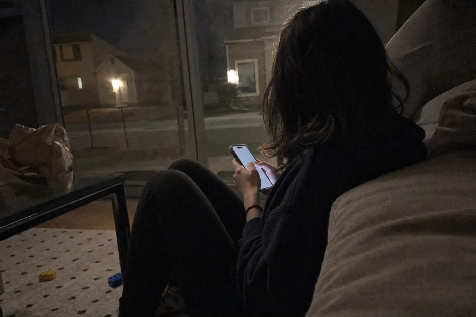 A woman sitting on a living room floor at night, seen from behind, lit by her phone