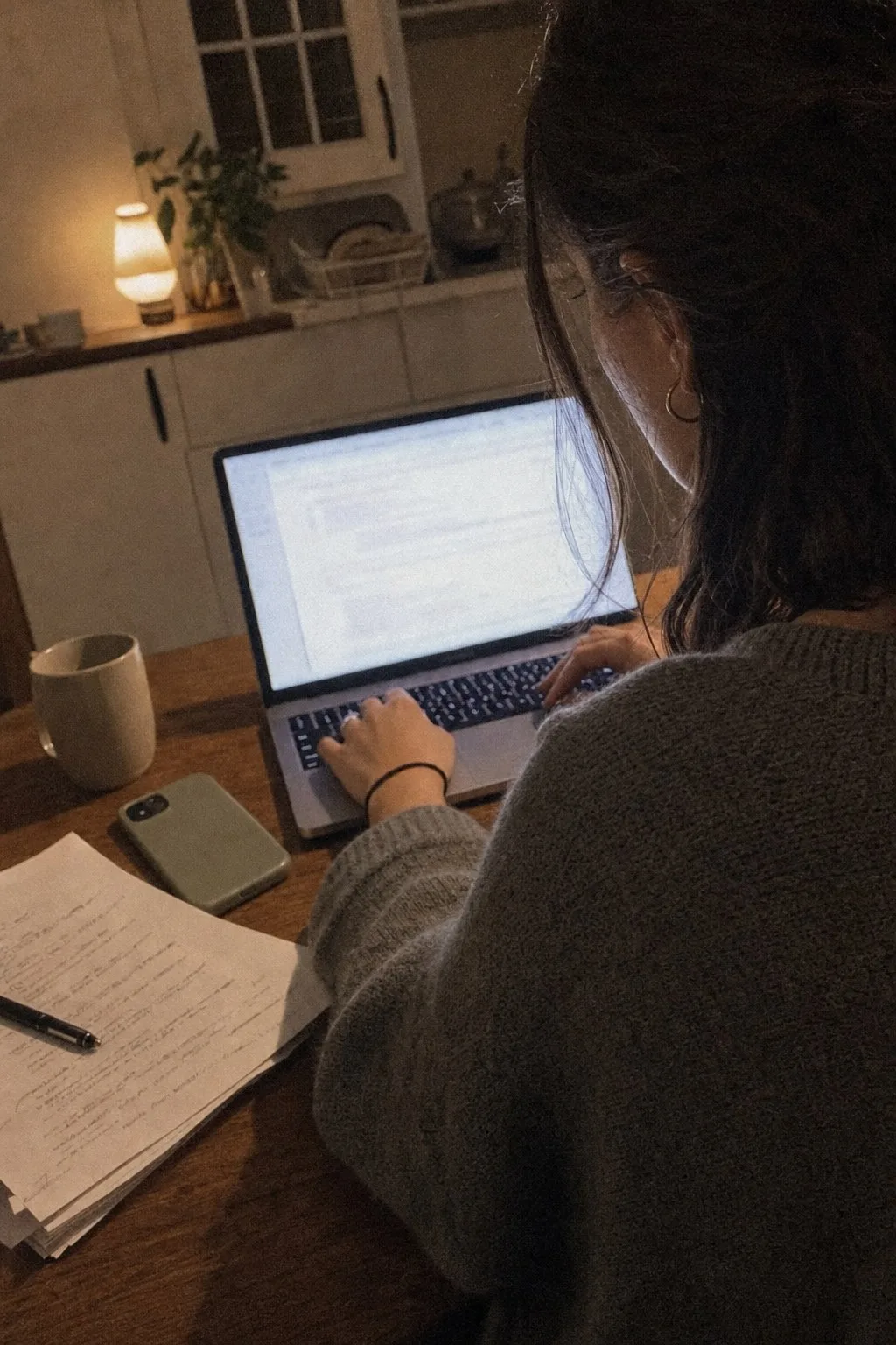 Over-the-shoulder view of someone typing on a laptop at night beside a stack of marked-up pages