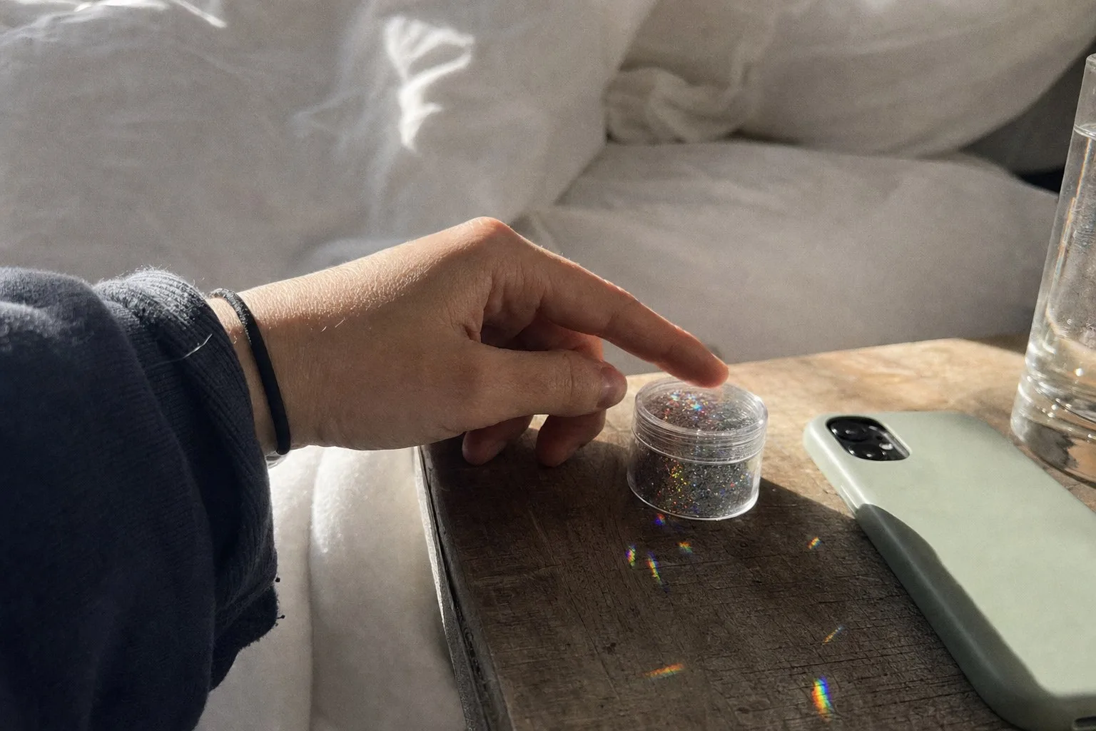 A fingertip resting on the lid of a small jar of holographic glitter on a bedside table
