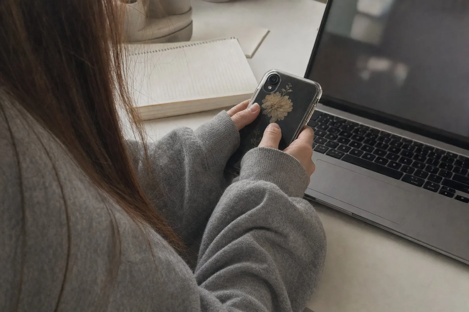 Hands holding a phone on a desk beside a laptop and notebook