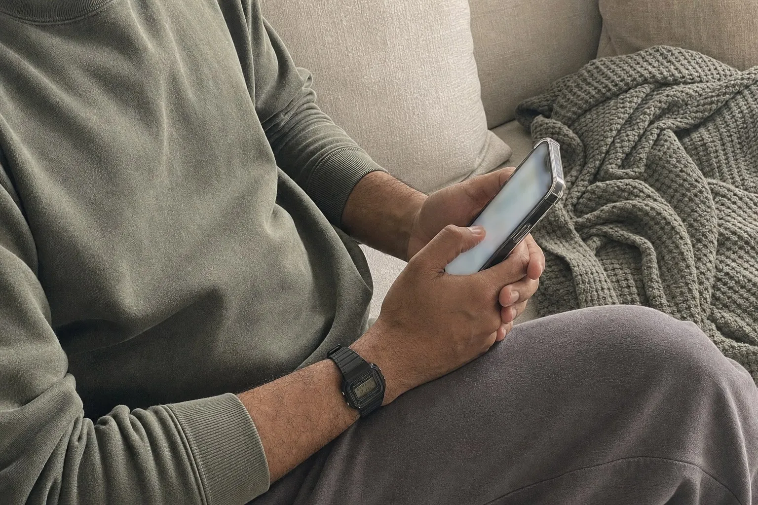 A man’s hands holding a phone on his knee while he sits on a sofa