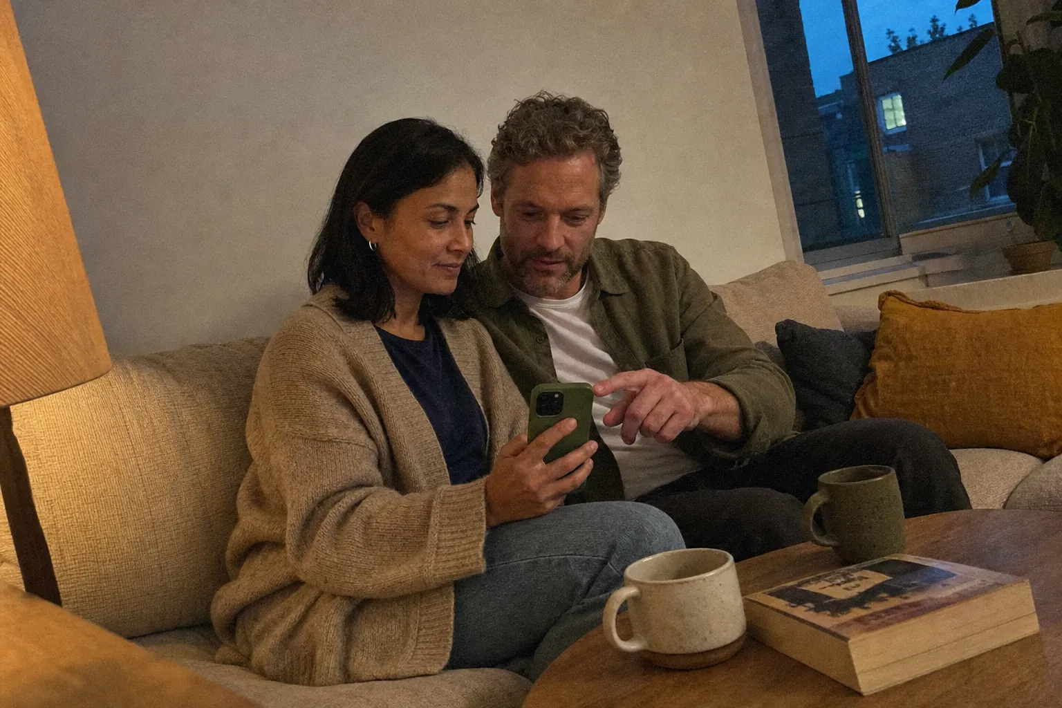 A couple on a sofa in the evening looking at one phone together while he points at the screen